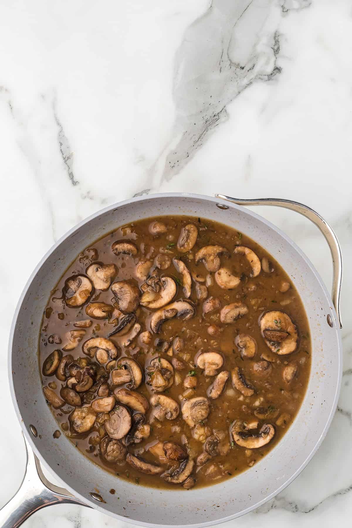A skillet filled with a brown mushroom sauce, sitting on a white marble surface.