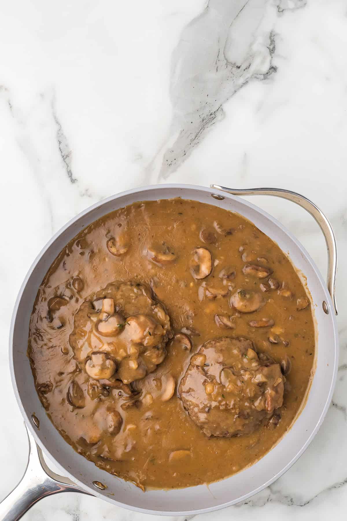 A skillet with two Salisbury steaks in mushroom gravy on a white marble countertop.