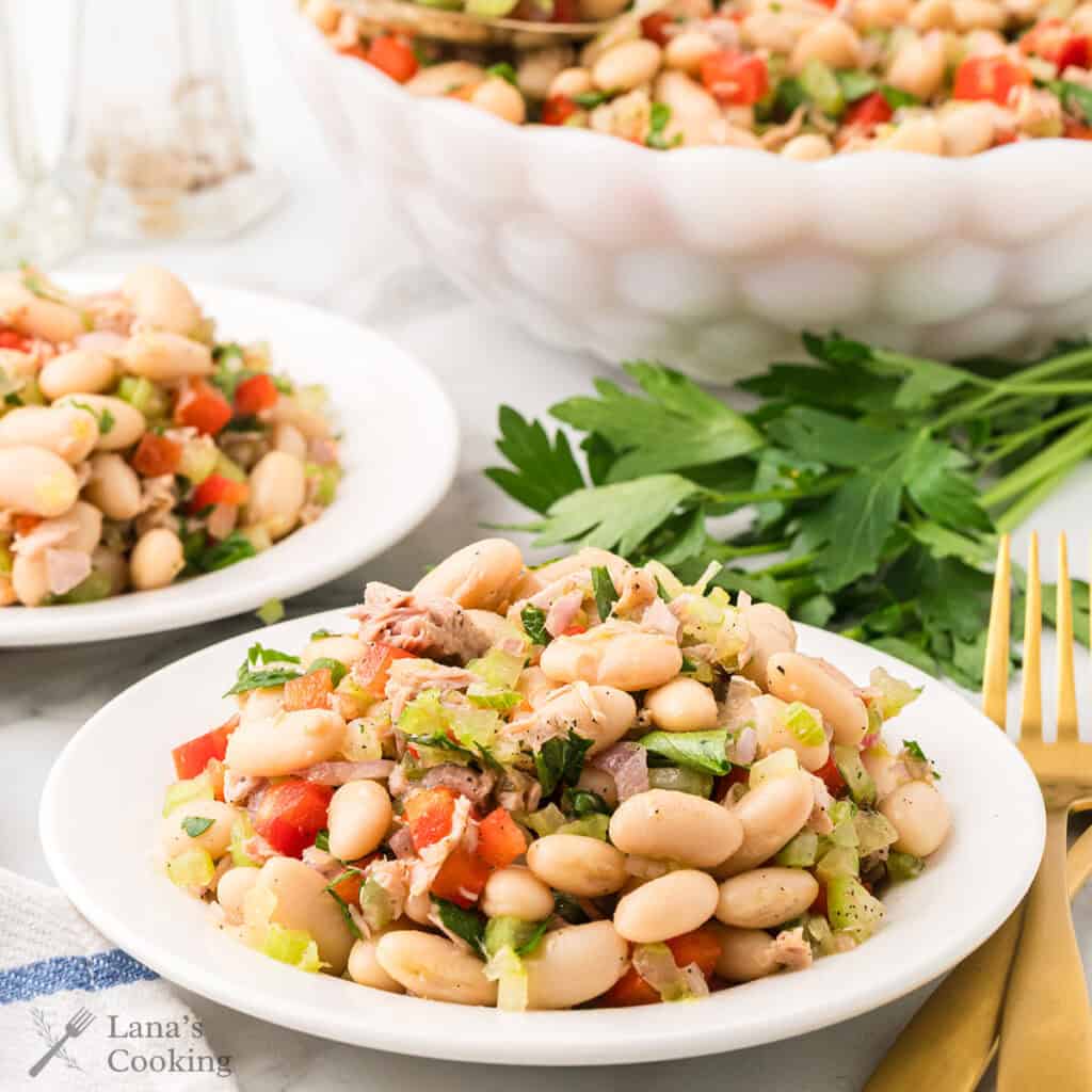 A white bowl and plates filled with white bean tuna salad with red bell pepper, celery, and herbs on a table.