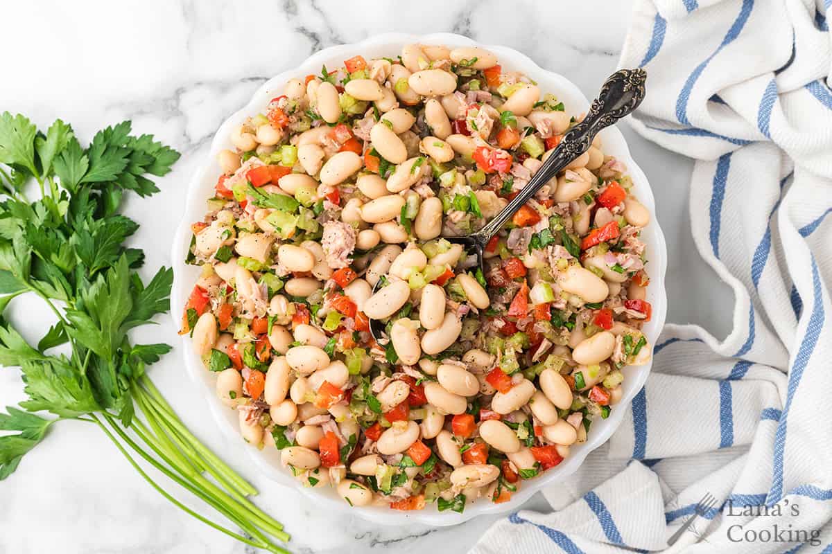 A bowl of white bean tuna salad with vegetables and herbs, with a spoon, next to fresh parsley on a striped cloth.