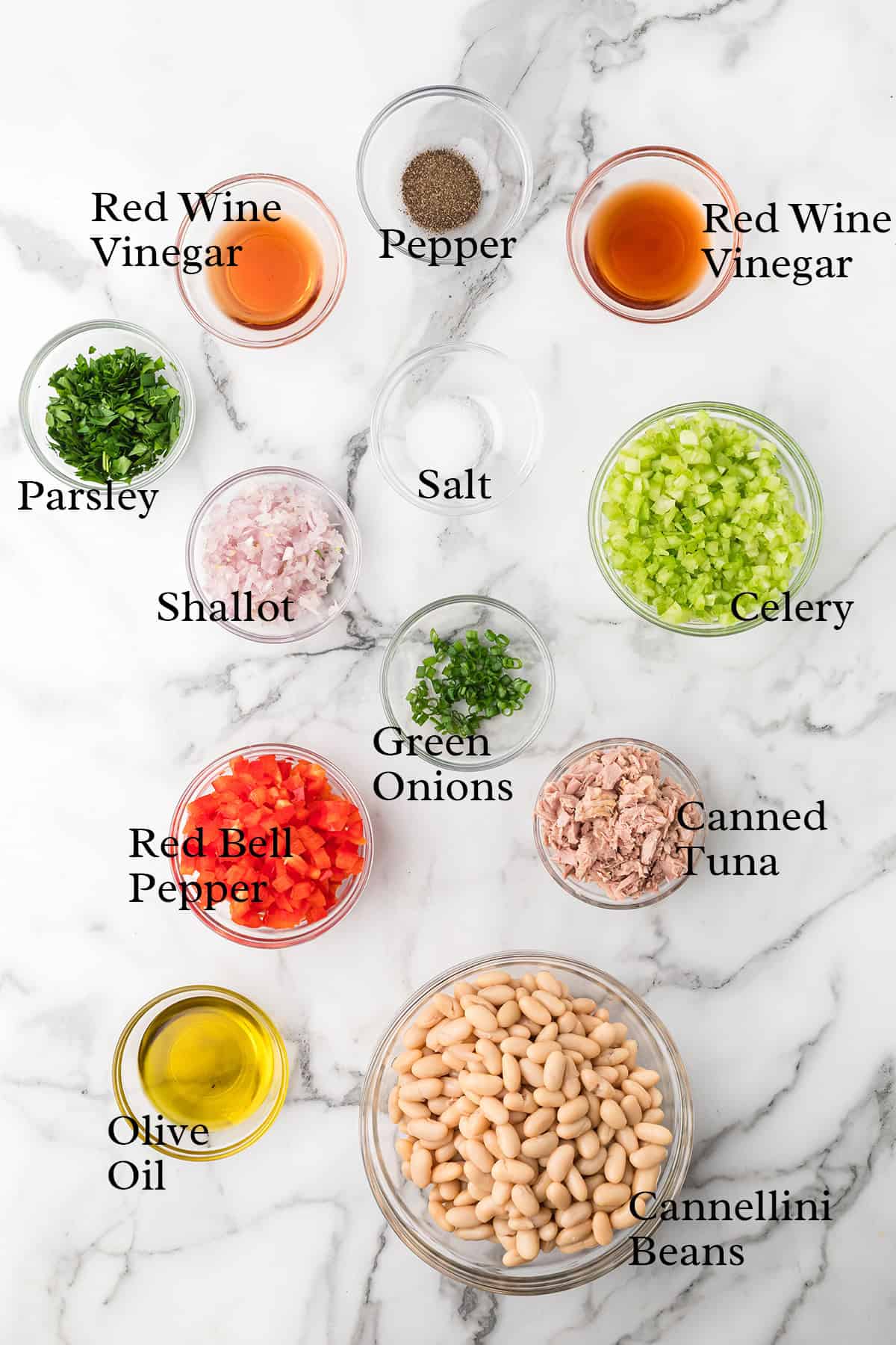 Ingredients for a tuna and bean salad arranged in bowls on a marble surface, each labeled with its name.