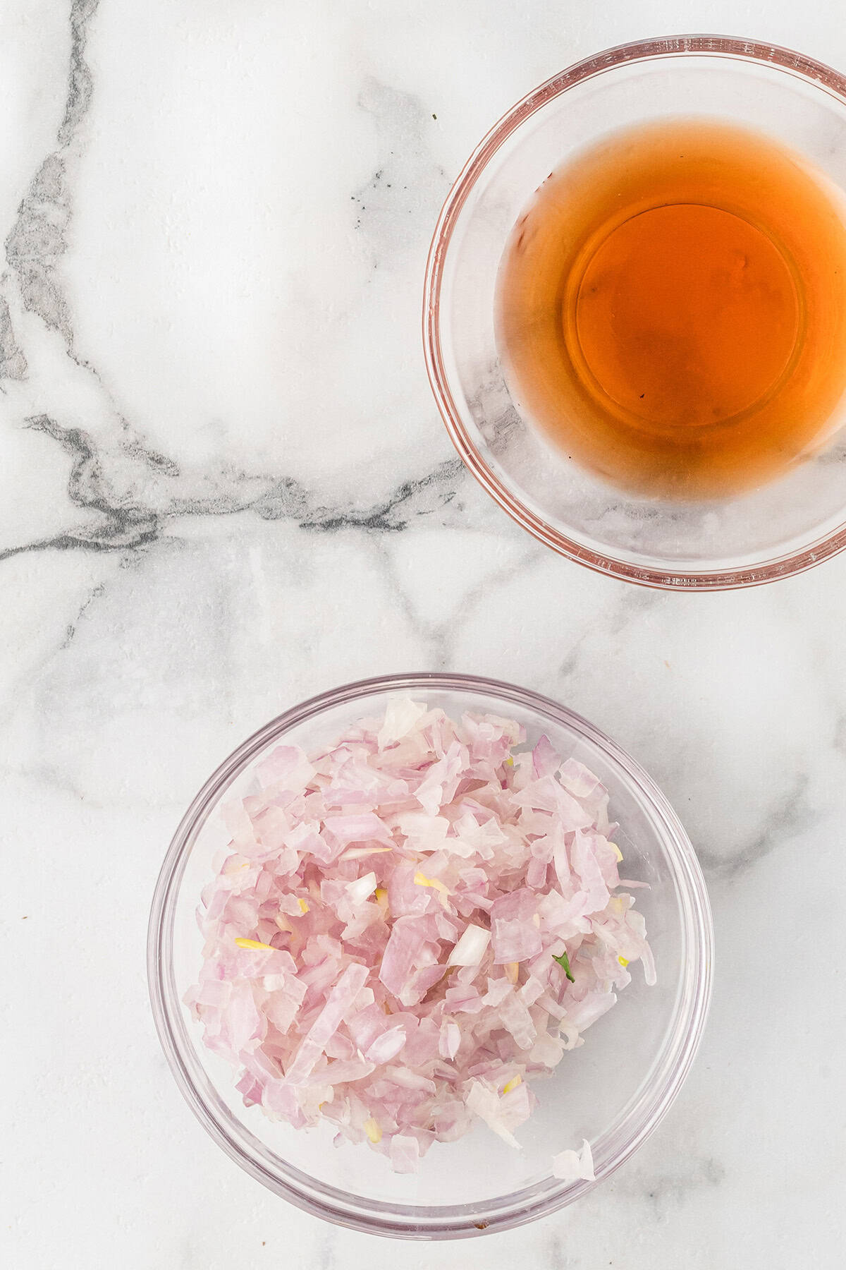 Two glass bowls on a marble surface, one with chopped shallots and one with red wine vinegar.