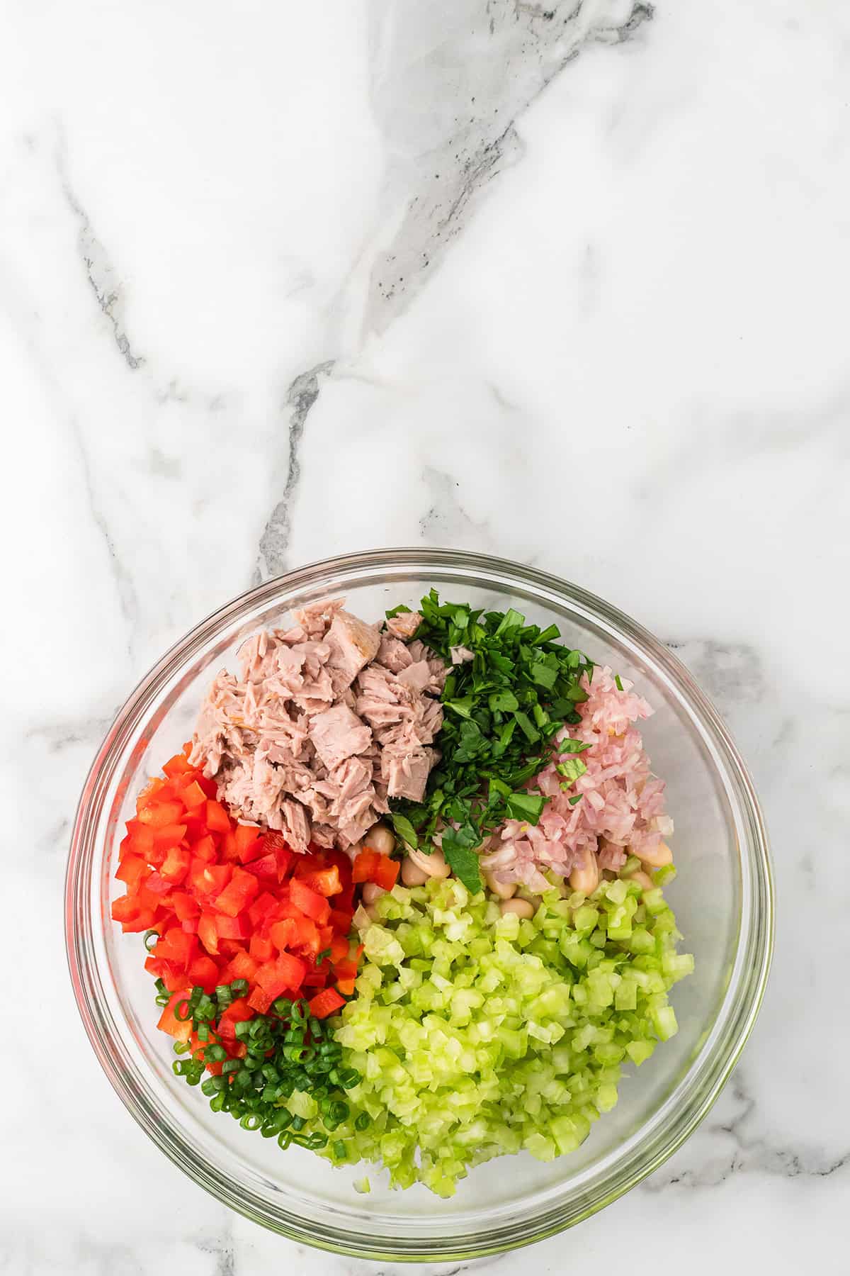 Glass bowl with tuna, chopped red bell pepper, celery, green onions, parsley, and shallots on a marble surface.