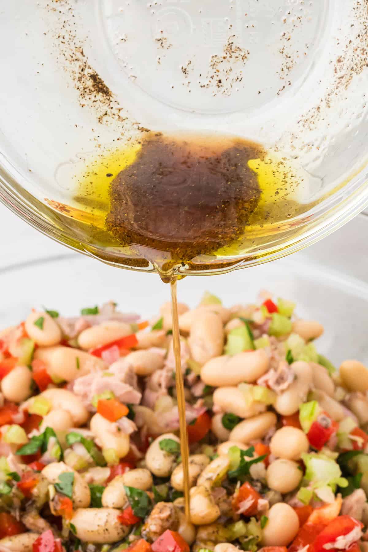 Olive oil dressing is being poured from a bowl over a bean salad with vegetables and herbs.