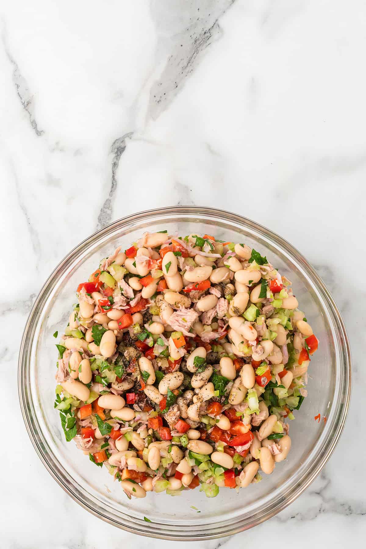 A glass bowl of bean and tuna salad with chopped vegetables and herbs on a marble surface.