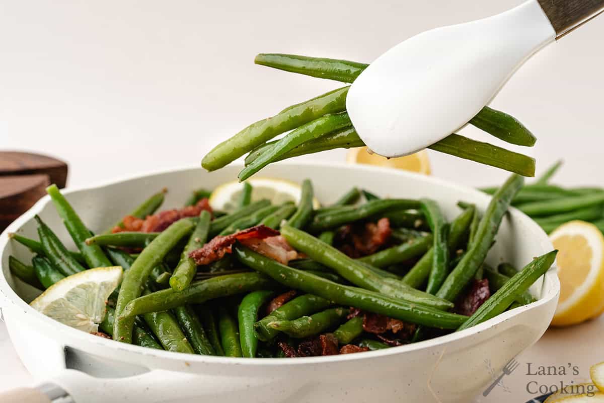 A white pan of green beans with bacon and lemon slices; tongs hold several green beans above the pan.