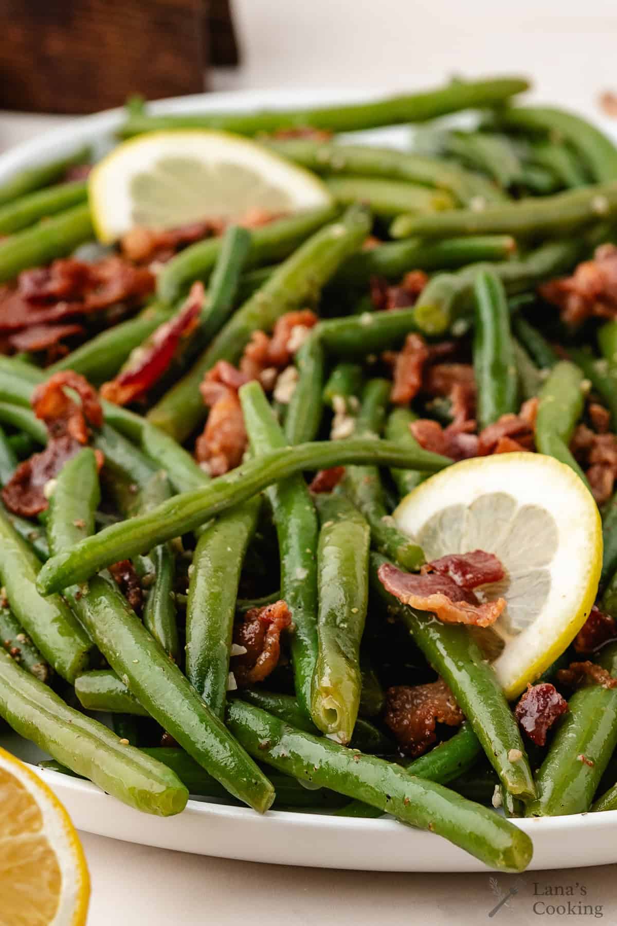 Green beans mixed with crispy bacon pieces, garnished with lemon slices, served on a white plate.