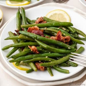 Green beans sautéed with bacon on a white plate, garnished with lemon slices, and a fork beside the food.