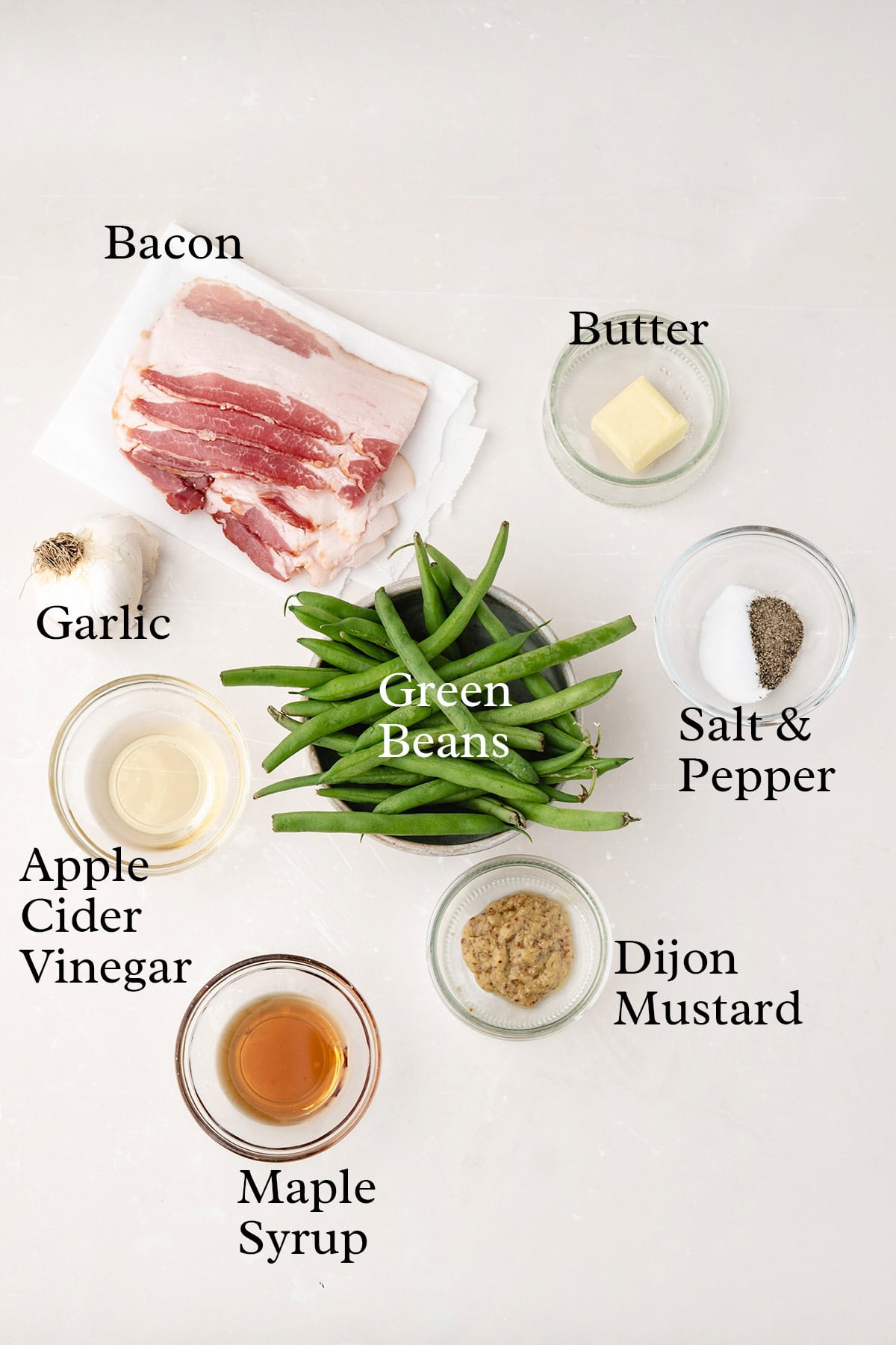 Ingredients on a white surface: bacon, green beans, butter, garlic, salt & pepper, apple cider vinegar, maple syrup, Dijon mustard.