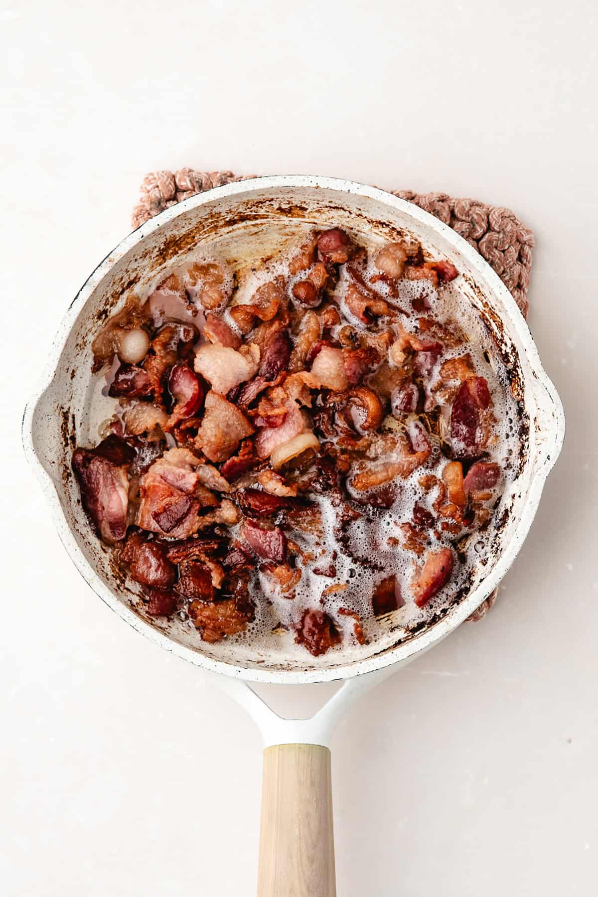 Crispy bacon pieces cooking in a white skillet with rendered fat on a light background.