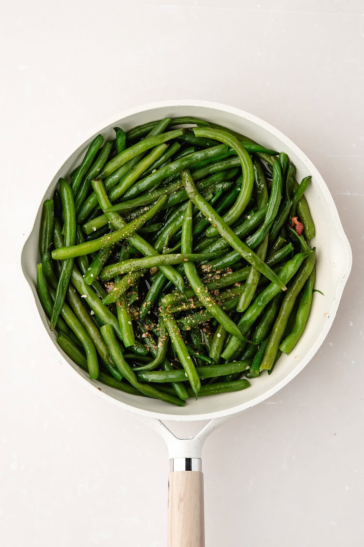 White skillet filled with cooked green beans, sprinkled with seasoning, on a light background.