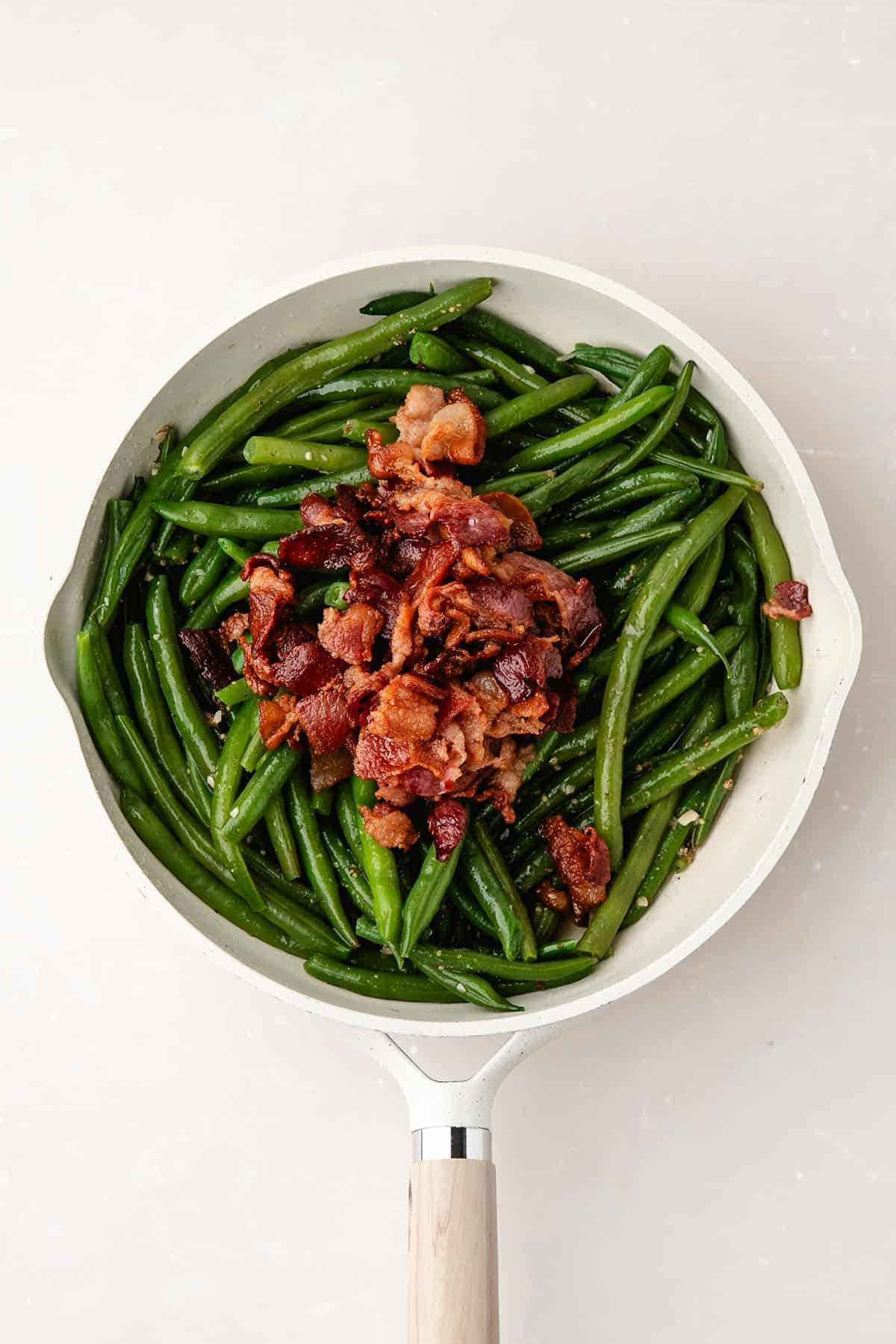 A white skillet with cooked green beans topped with crispy bacon pieces on a light surface.