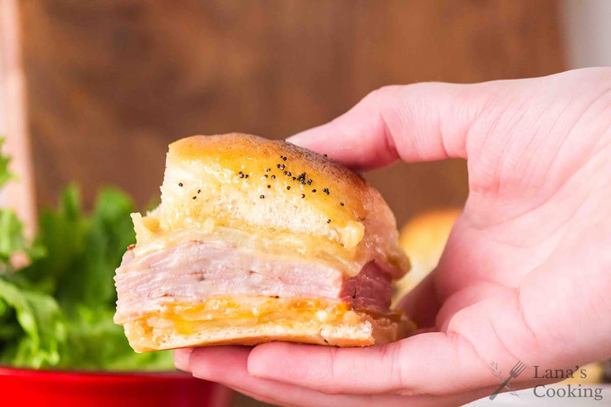 A hand holds a ham and cheese slider on a bun, with lettuce and a red bowl blurred in the background.