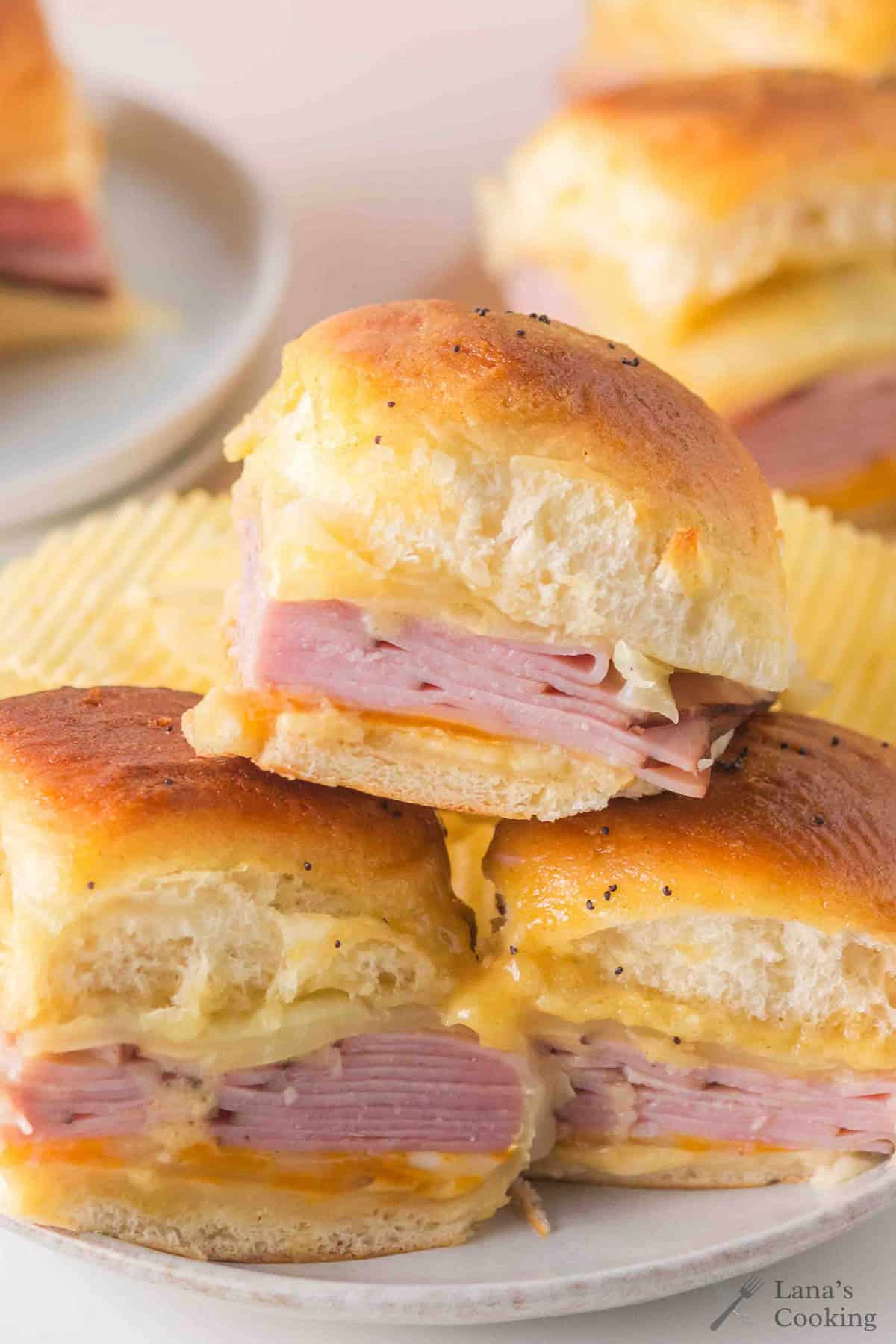 Stacked ham and cheese sliders on soft buns, with melted cheese and potato chips in the background.