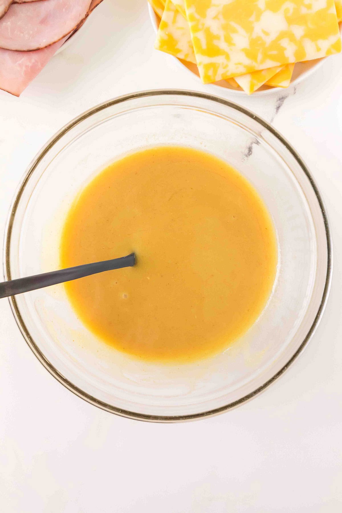 Glass bowl of honey mustard sauce with a spoon, next to slices of ham and marbled cheese on a white surface.