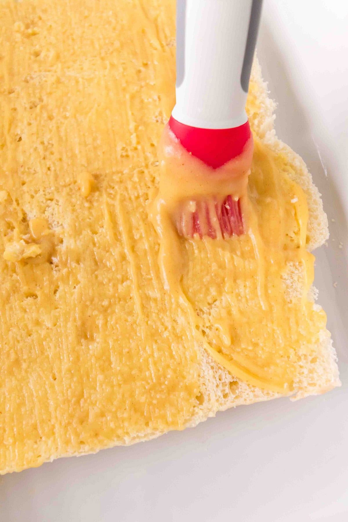 A red pastry brush spreads butter-mustard-honey mixture over a sheet of dough on a white surface.