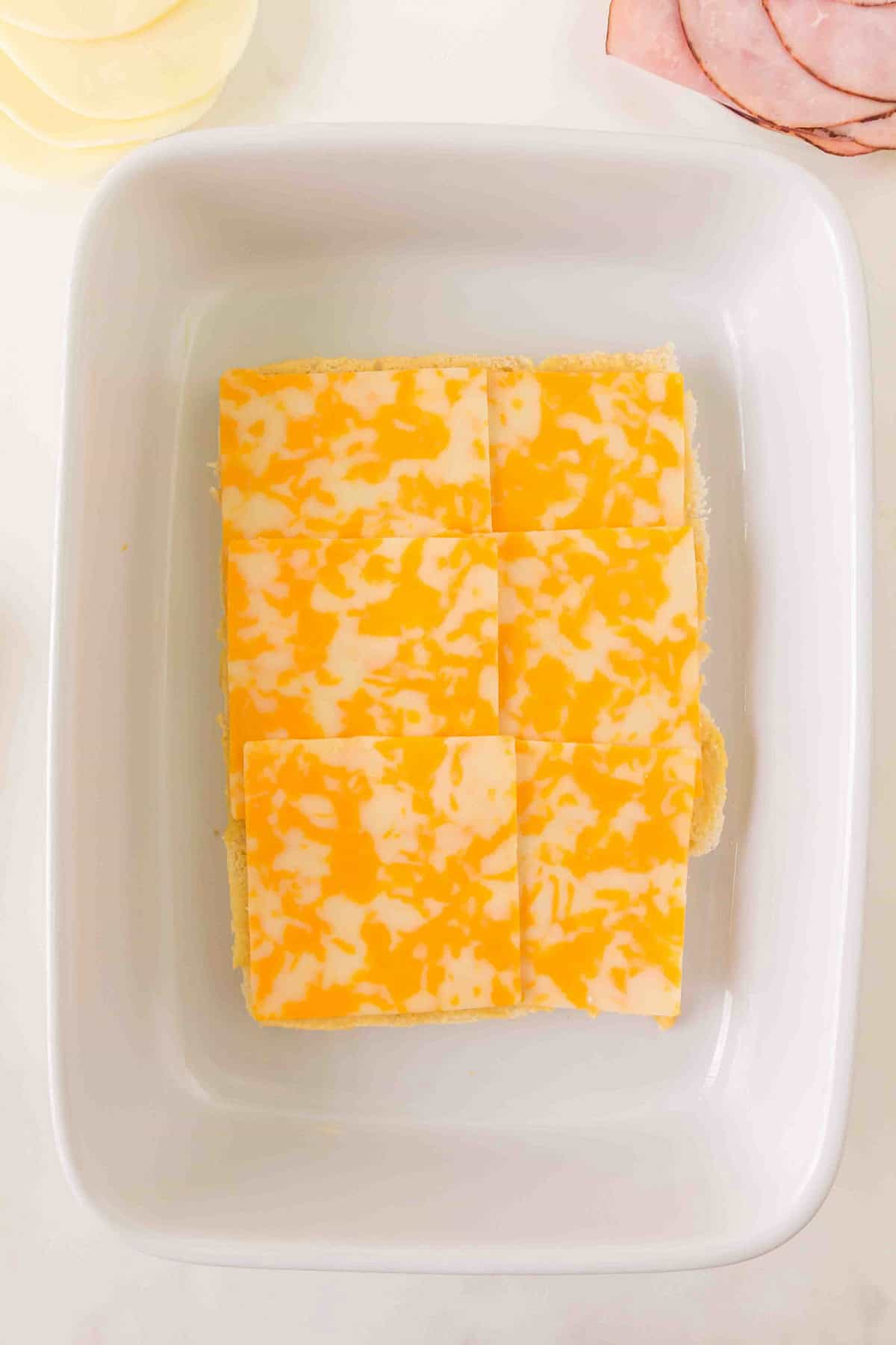 Six slices of Colby Jack cheese on the bottom of slider rolls in a white baking dish, viewed from above.