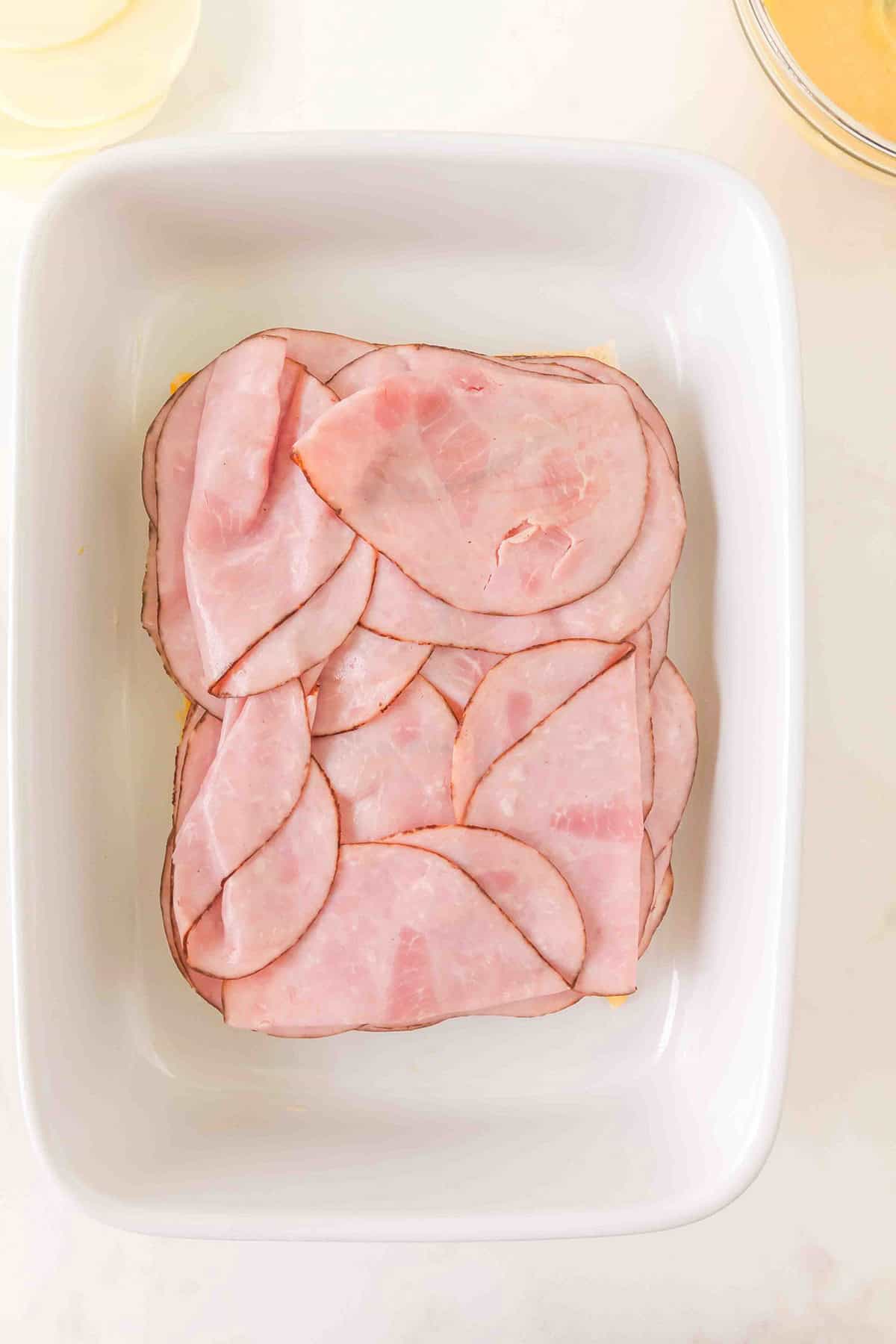 White dish with neatly layered slices of pink deli ham on a light surface, viewed from above.