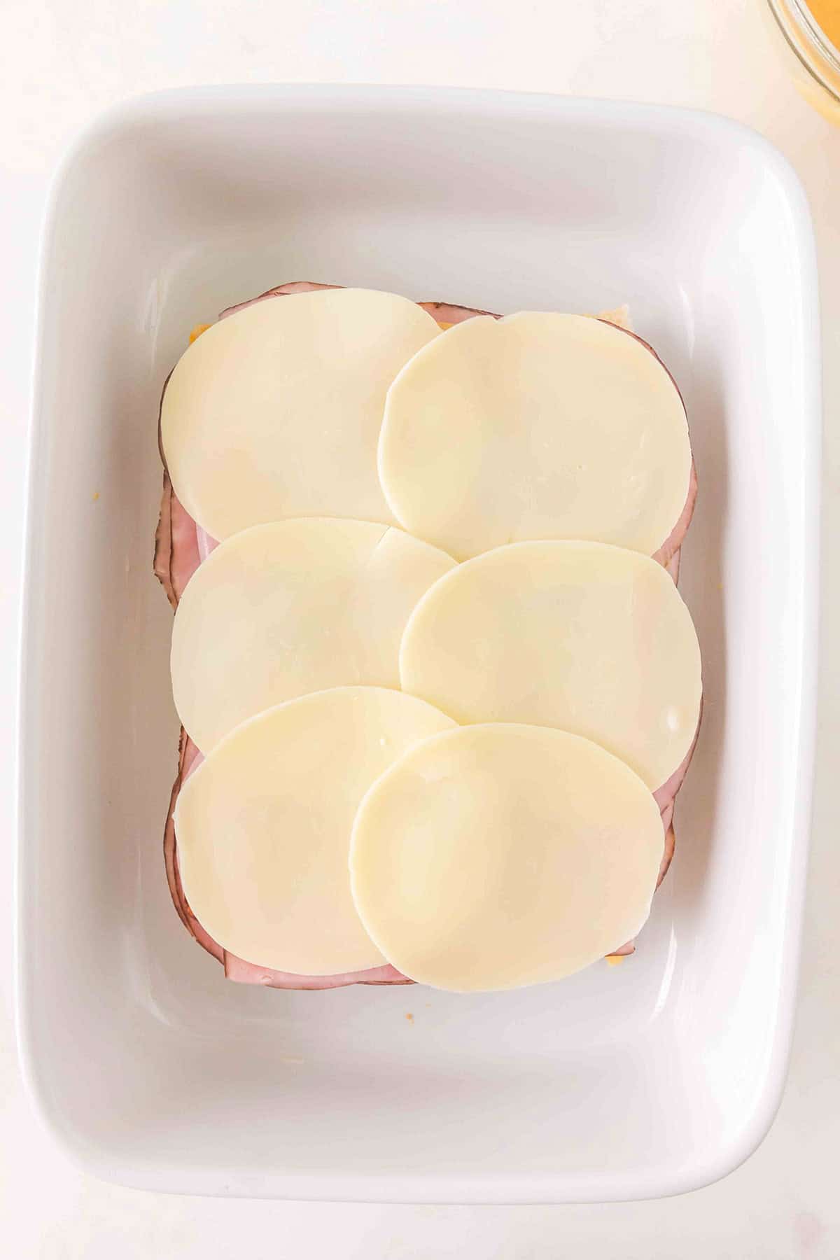 Six slices of provolone cheese on top of ham in a white baking dish, viewed from above.